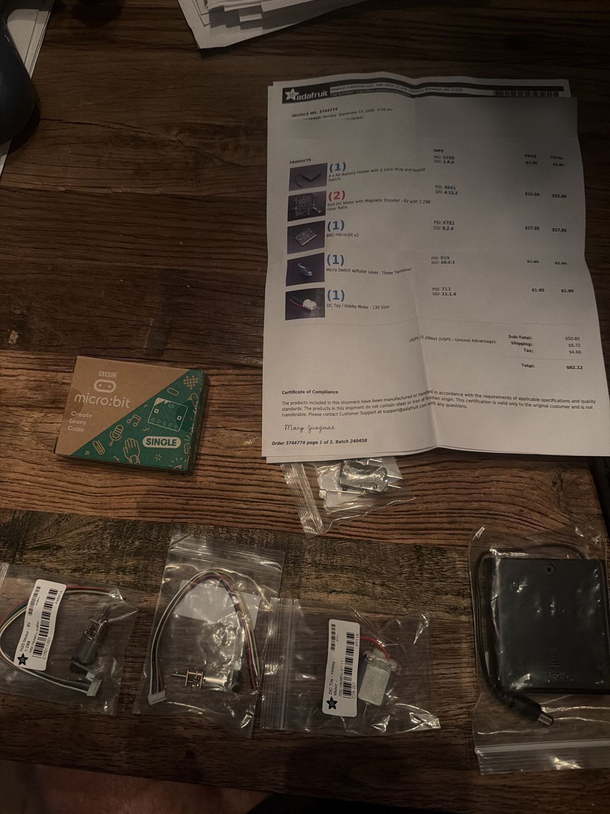 An Adafruit packing slip on a wooden table with a boxed micro:bit v2, two bagged N20 gear motors, a bagged microswitch, a bagged small hobby motor, and a black 4 AA battery holder