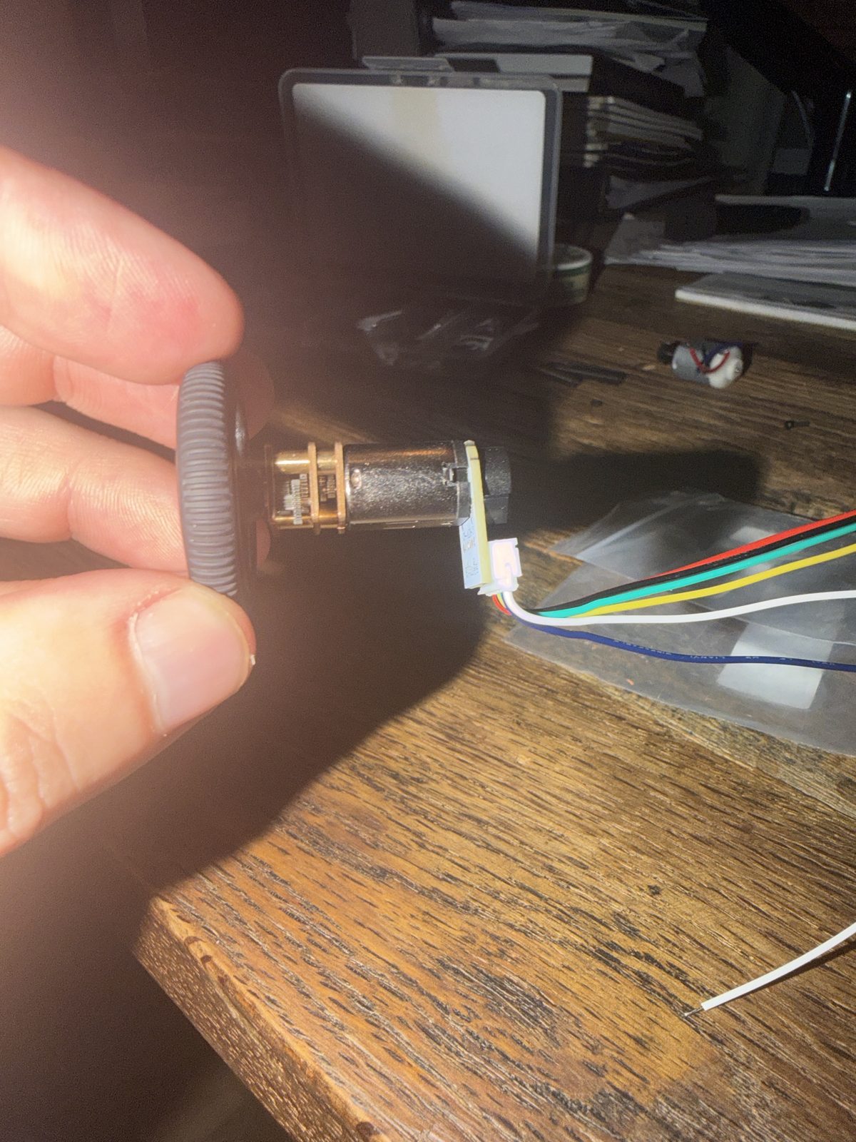 A hand holding an N20 gear motor by its black wheel, with a small circuit board on the back end and a white plug hanging off one side, six colored wires running away across a wooden table