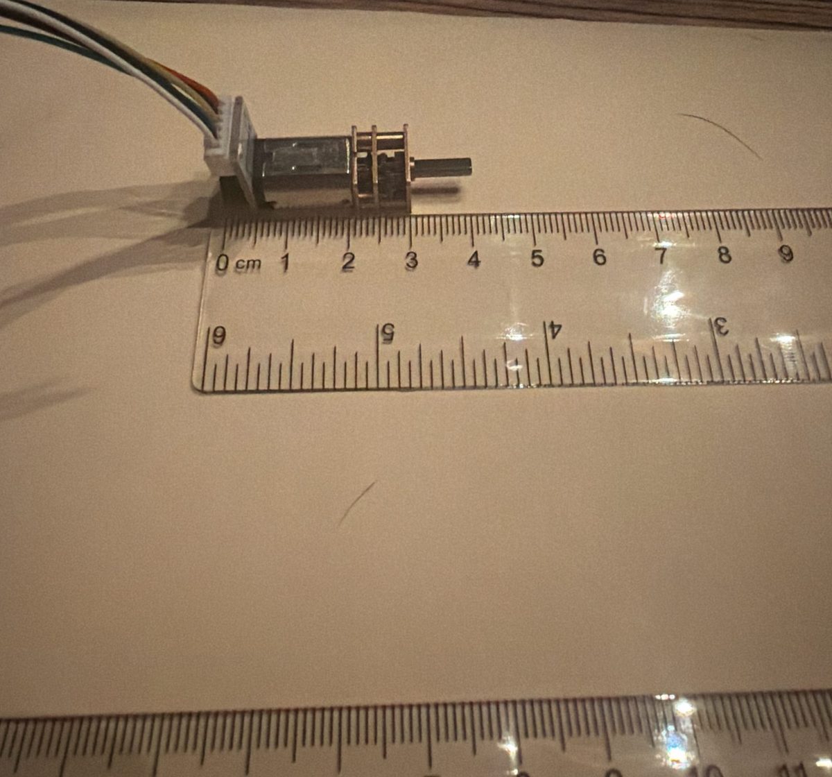 An N20 gear motor lying on its side on paper next to a clear centimeter ruler, the plug end at 0 and the front of the gearbox at about 3.3 cm, with the shaft sticking out past it