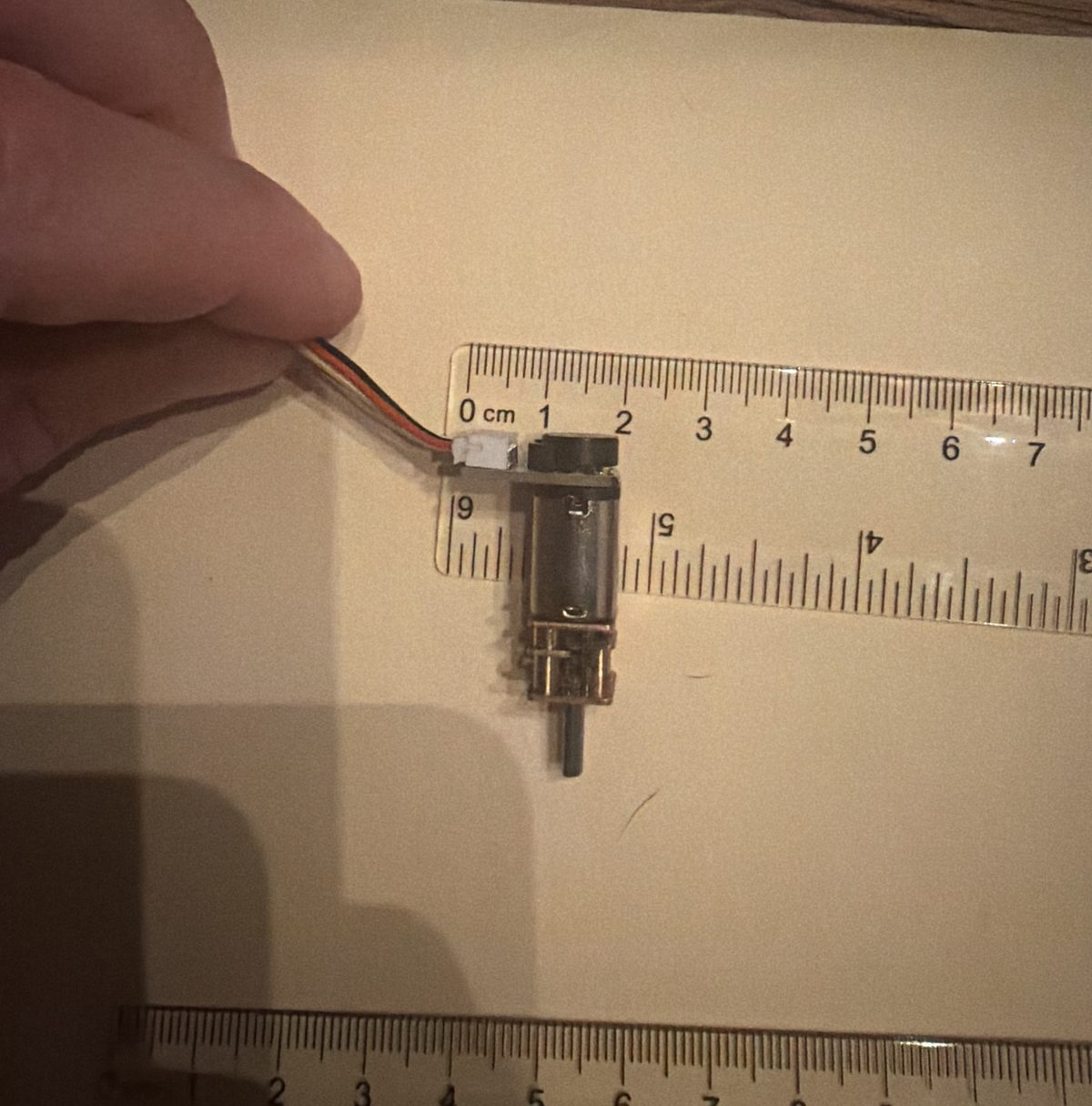 An N20 gear motor standing on paper with a clear centimeter ruler across its back end, the encoder board and white plug reaching past the motor body to the left