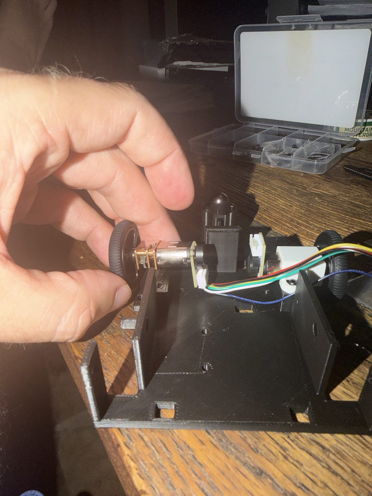 A hand holding an N20 gear motor with a wheel over a black 3D printed robot base, the small green encoder board and white plug on the back of the motor sitting against the base plate, a second motor with colored wires already in a white bracket on the far side