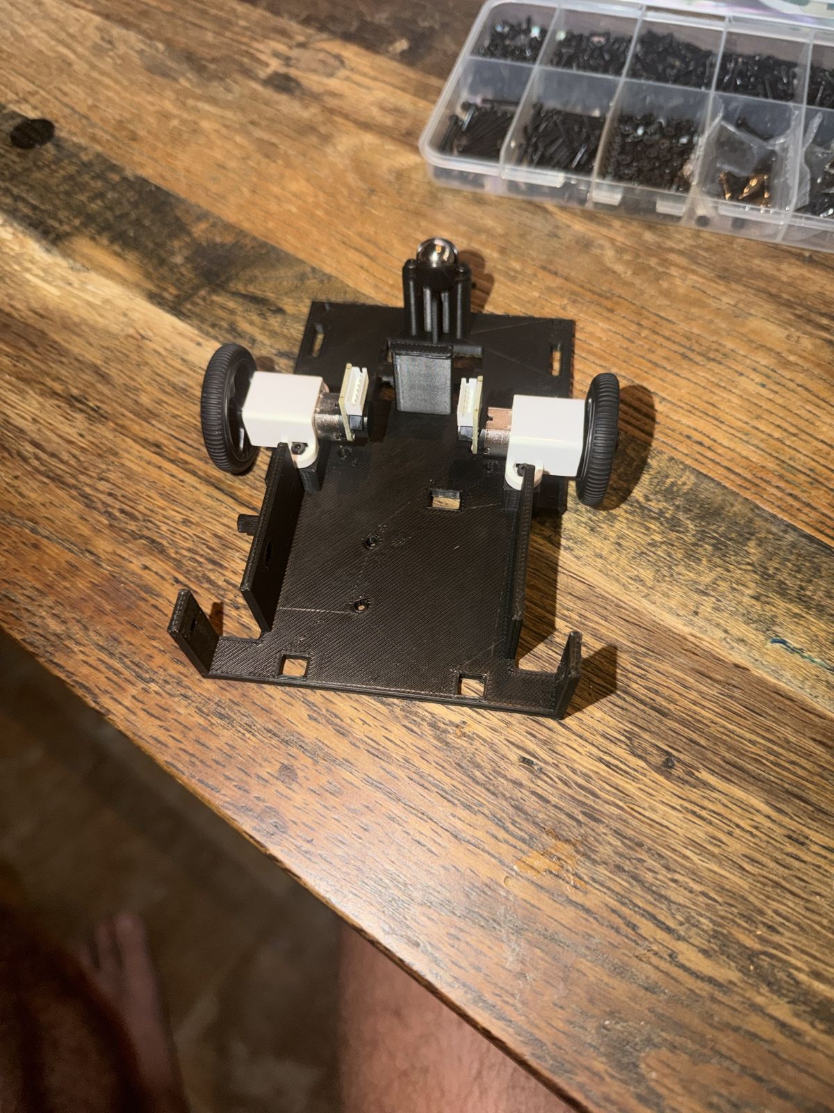 A black 3D printed robot base upside down on a wooden table, with two small gear motors in white brackets, a black rubber wheel on each side, a metal ball caster on a post, and a box of black screws behind it