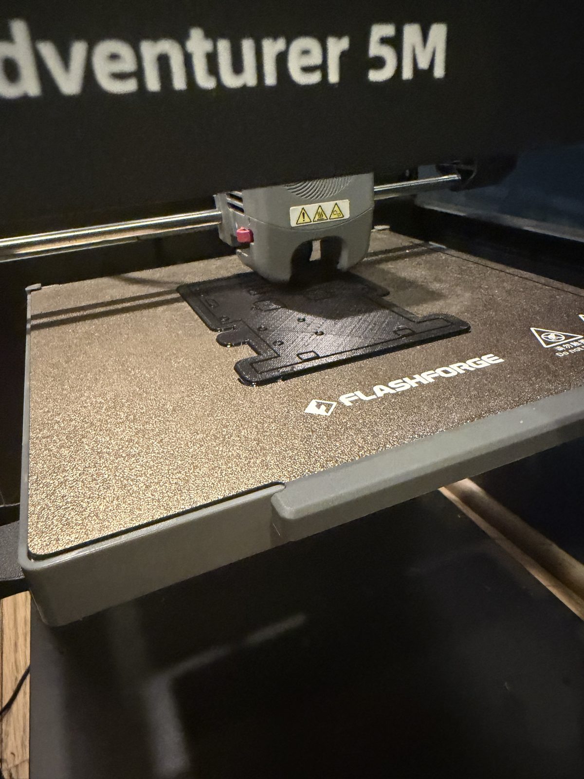 Close up of a 3D printer nozzle over a thin black robot base outline on a textured Flashforge build plate, with holes and cutouts visible in the first layers