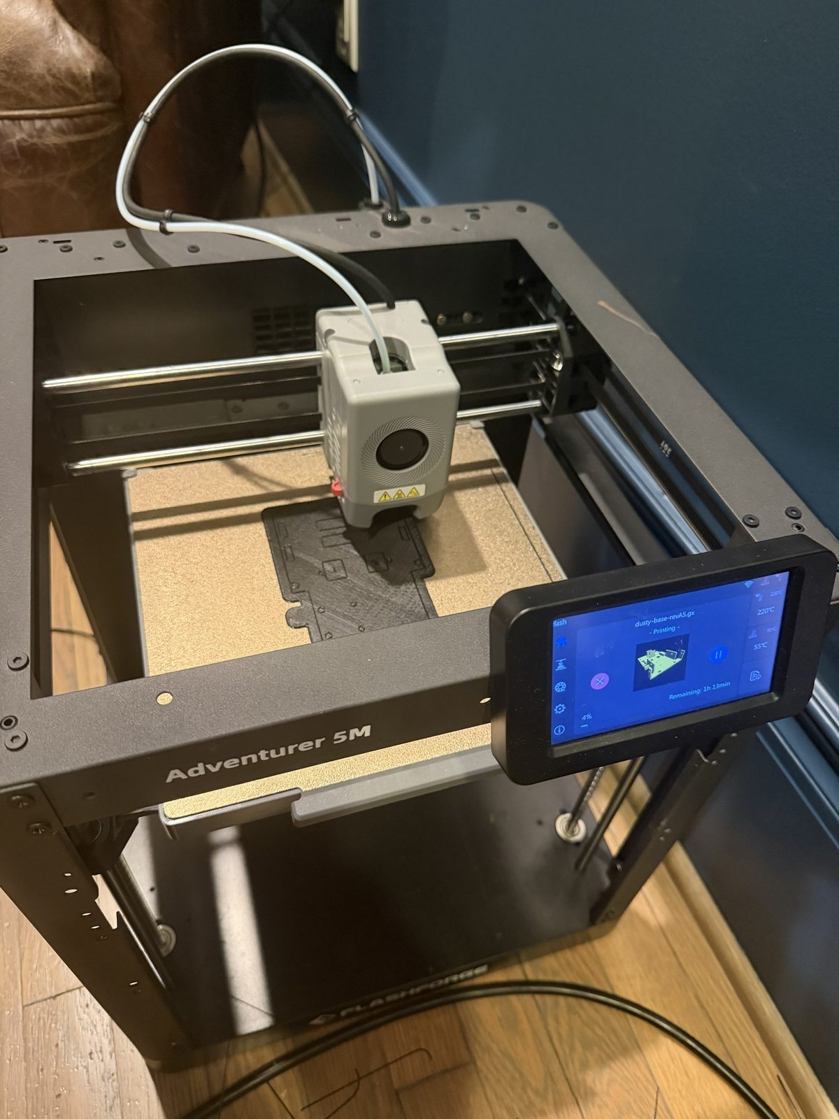 A Flashforge Adventurer 5M 3D printer from above, printing a flat black robot base on a gold textured build plate, with the touch screen showing dusty-base-revA5 printing at 4 percent and 1 hour 13 minutes remaining