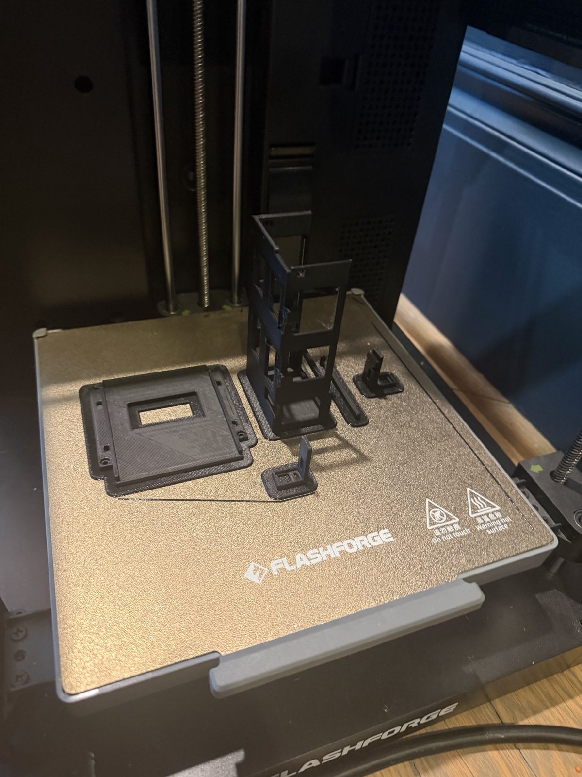 A Flashforge 3D printer build plate holding black printed parts: a flat deck with a square window, a tall open box standing on end, a thin bar, and two small L-shaped arms
