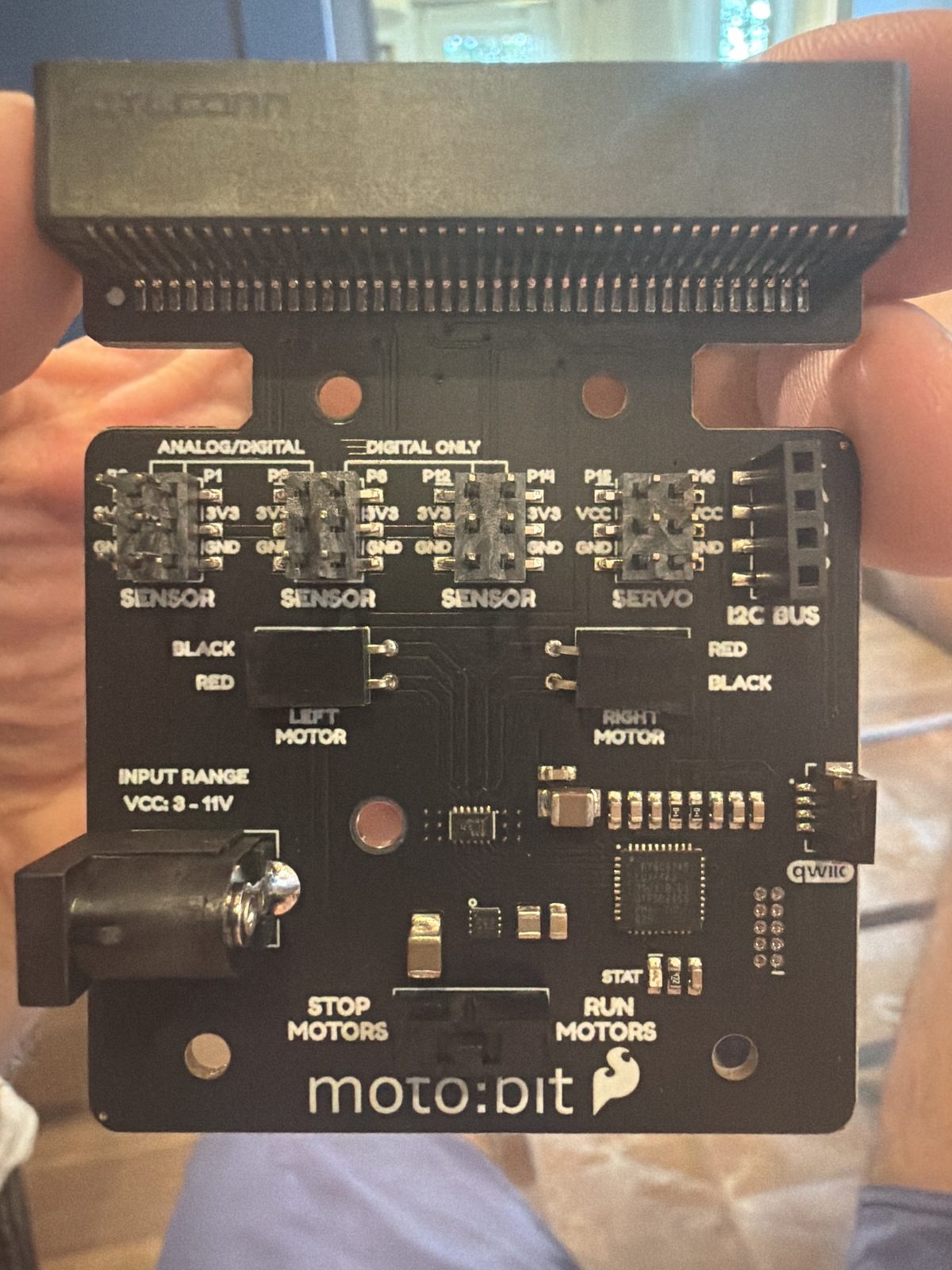 Close up of a black SparkFun moto:bit circuit board held in two hands, showing the micro:bit edge connector slot, sensor and servo pin headers, left and right motor connectors, a barrel power jack, and a stop motors / run motors switch
