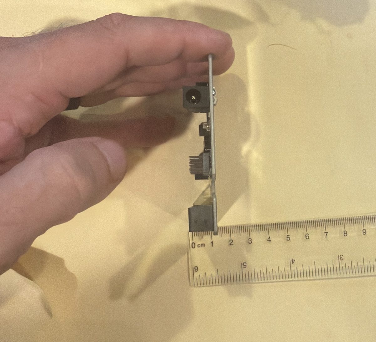 A hand holding a moto:bit board edge-on above a clear ruler on yellow paper, showing the barrel jack, pin headers and micro:bit slot sticking out from one side of the board