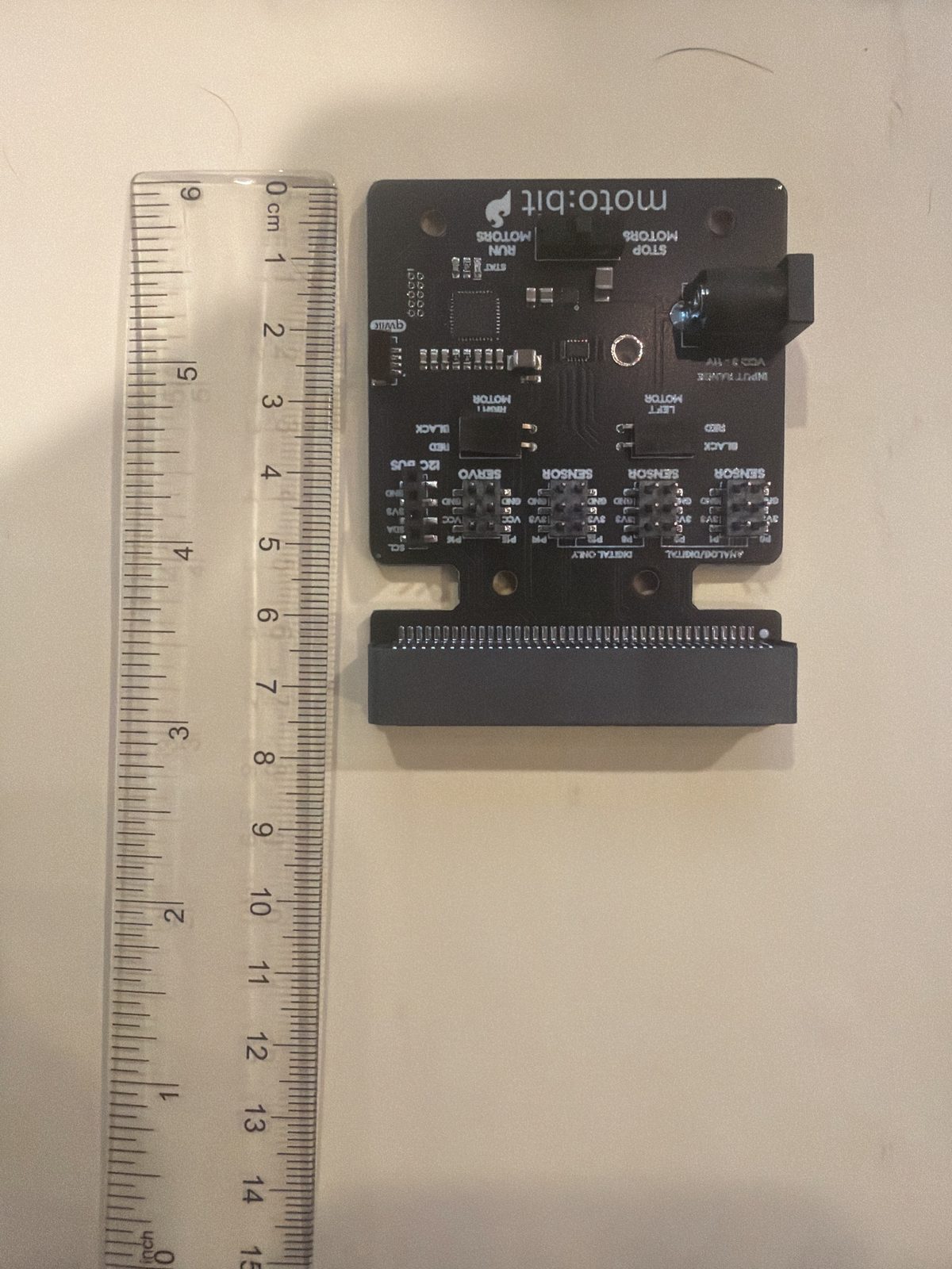 A moto:bit board lying flat on paper next to a clear ruler running top to bottom, the board reaching from 0 to about 7.6 centimeters