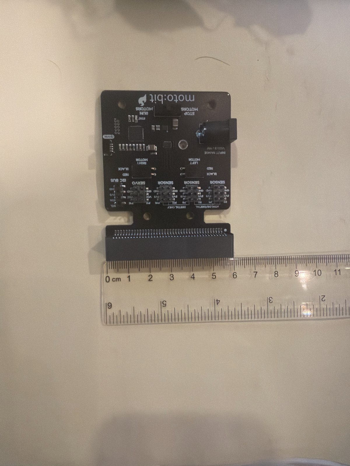 A moto:bit board lying flat on paper with a clear ruler along the bottom edge of its micro:bit connector, the connector spanning about 6 centimeters