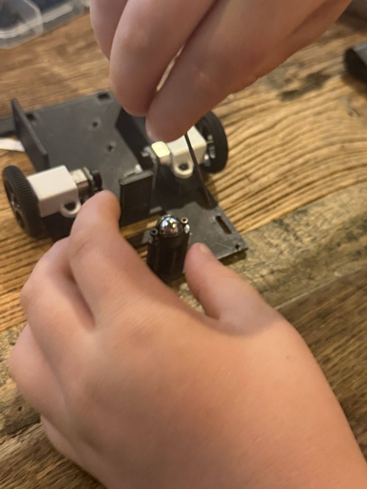 A boy's hands on a black 3D printed robot base on a wooden table, one hand holding a metal ball caster on a black post while another hand turns a small hex key, with a wheel and white motor bracket on each side