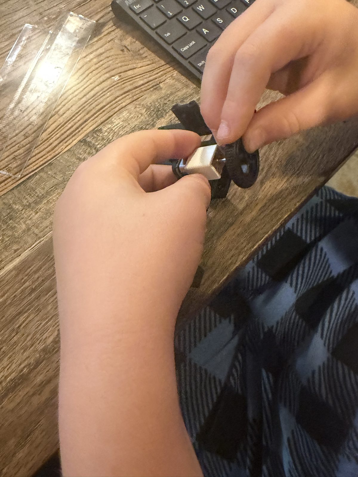 A boy's hands at a wooden desk pressing a black rubber wheel onto a small gear motor held in a white bracket on a black 3D printed robot base, a keyboard and a clear ruler nearby