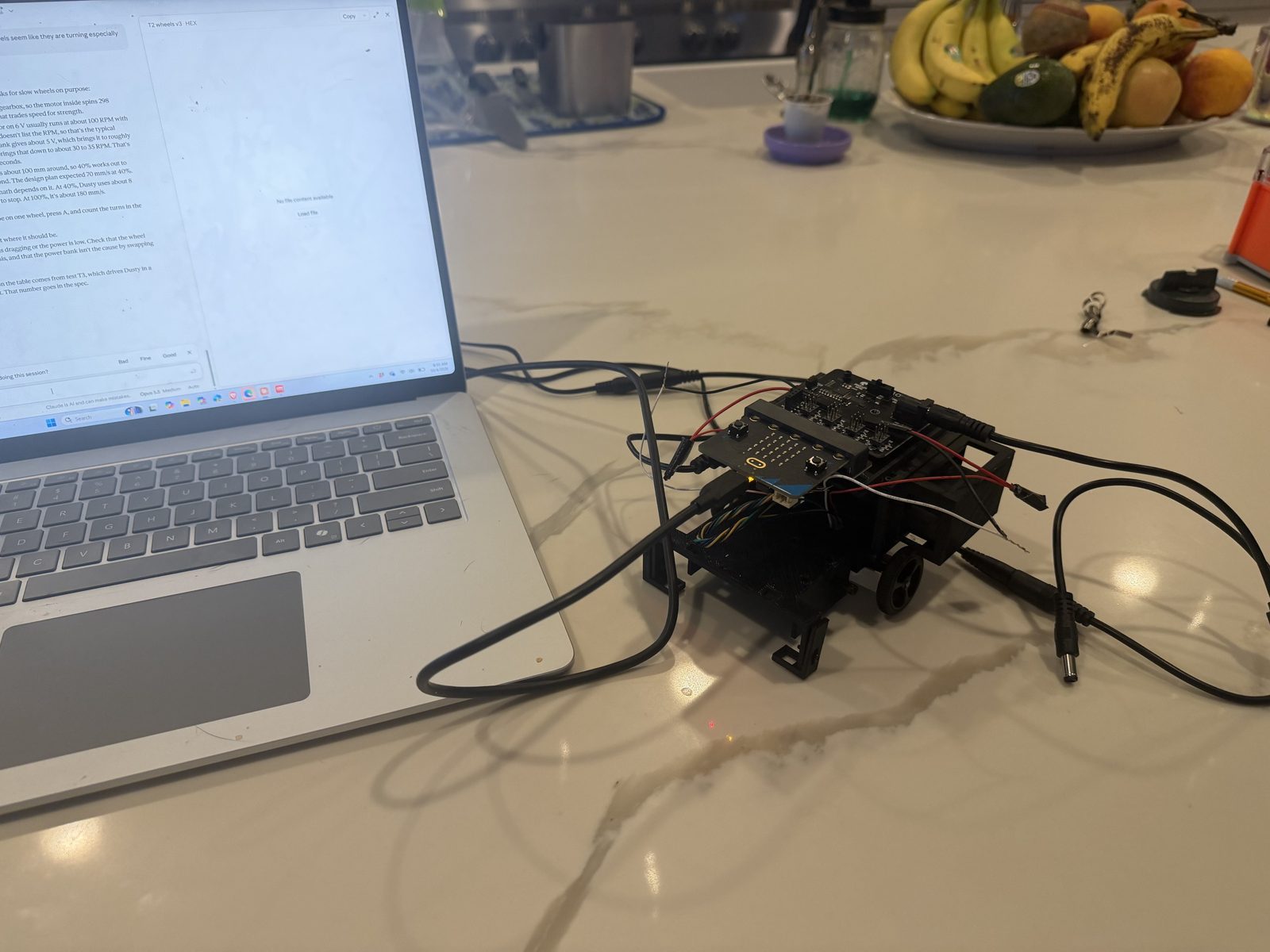 A laptop on a kitchen counter with a USB cable running to the robot, the micro:bit showing a small yellow light on its back