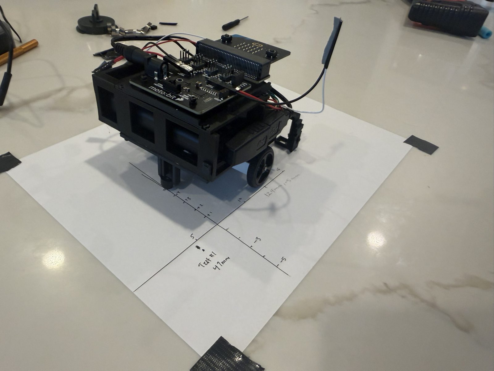The robot sitting on a taped sheet of white paper with hand-drawn x and y axes, tick marks in centimeters, and pencil notes reading Test 1 47mm and 124mm, -5mm
